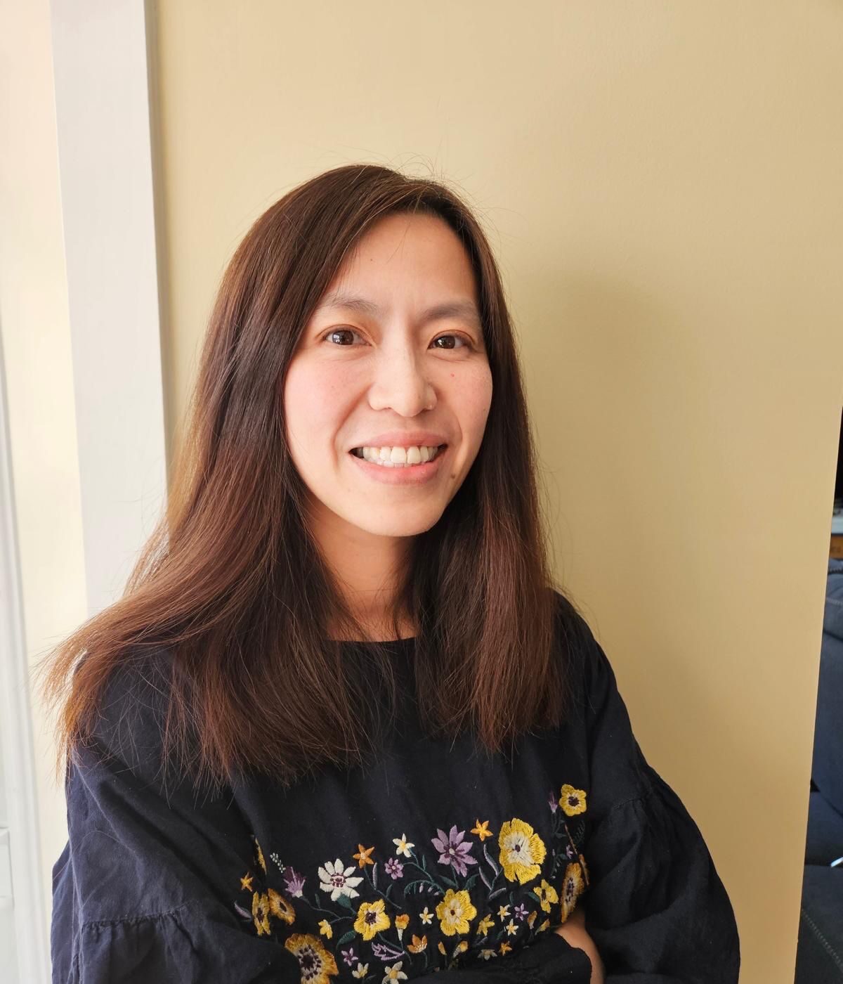 A woman with long, straight brown hair is smiling warmly at the camera. She is wearing a black blouse with colorful floral embroidery and is standing against a light yellow wall.